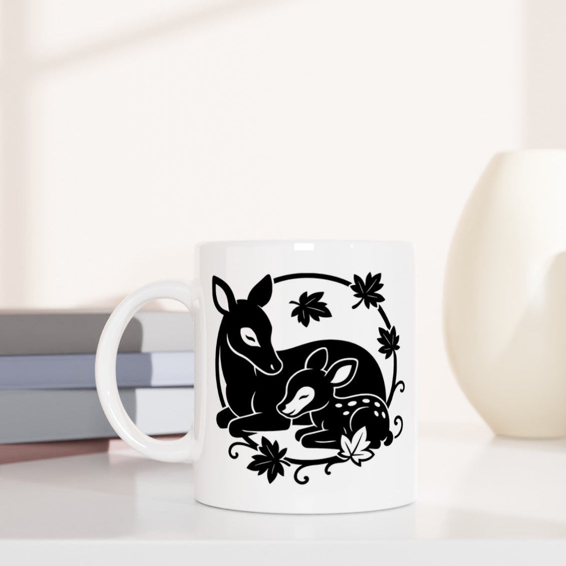 Weiße Tasse mit schwarzem Motiv von zwei Rehen und Blättern, daneben gestapelte Bücher und zwei Vasen in Beige- und Cremetönen.