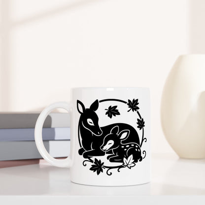 Weiße Tasse mit schwarzem Motiv von zwei Rehen und Blättern, daneben gestapelte Bücher und zwei Vasen in Beige- und Cremetönen.